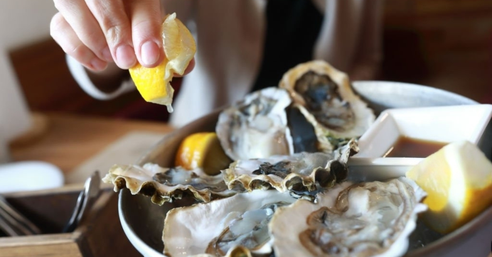 Image showing someone squeezing lemon juice on a half shell of East Coast oysters, with lemon wedges on the side.
